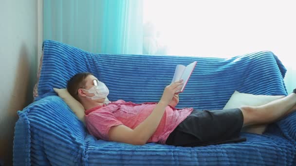young man reading a book at home in a mask from the coronavirus, self-isolation - Footage, Video