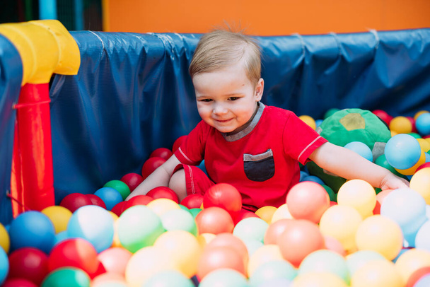 Happy laughing boy 1-2 years old having fun in ball pit on birthday party in kids amusement park and indoor play center. Child playing with colorful balls in playground ball pool. Activity toys for little kid. - Foto, afbeelding