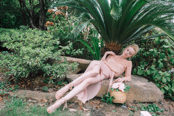 Чувственная молодая блондинка красивая милая и элегантная женщина в розовом сидя на скале и глядя в камеру над тропическими зелеными джунглями пальмового фона в Китае
. - Фото, изображение