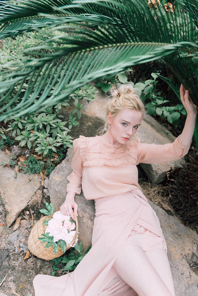 Чуттєва молода блондинка, красива і елегантна жінка, яка сидить на скелі в рожевому кольорі і і дивиться в камеру через тропічні зелені джунглі пальмовий фон у Китаї.. - Фото, зображення