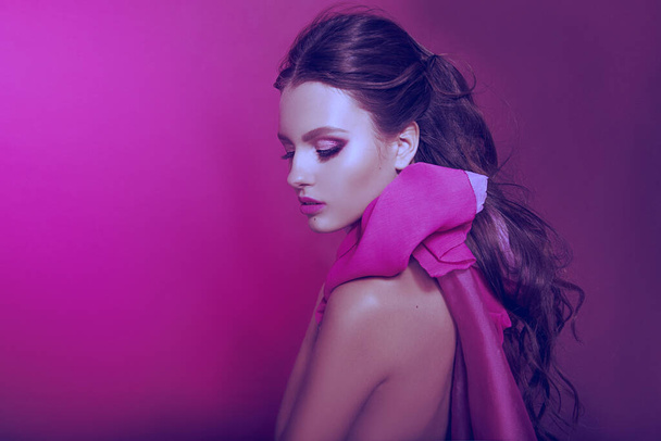 Mooi modieus meisje met lang krullend haar poserend met een roze strik. Het meisje in de studio op een roze achtergrond. Mode, schoonheid. Heldere kleren. Cosmetica voor haar, gezond haar. - Foto, afbeelding