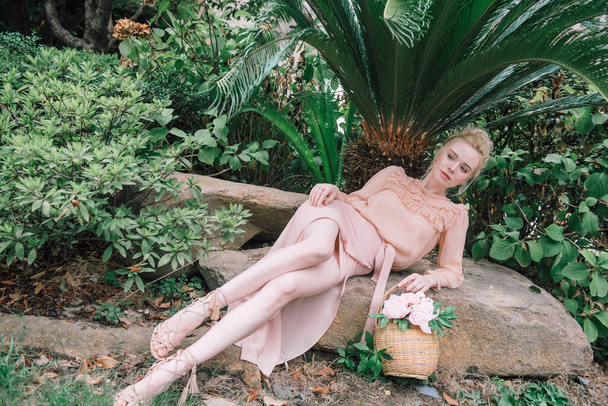 Sensueel jong blond mooi schattig en elegant vrouw dragen roze zitten op de rots en kijken in de camera over tropische groene jungle palm achtergrond in China. - Foto, afbeelding