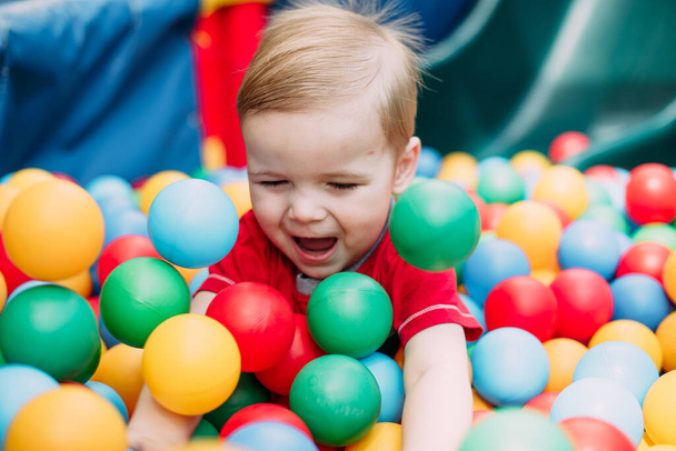 Happy laughing boy 1-2 years old having fun in ball pit on birthday party in kids amusement park and indoor play center. Child playing with colorful balls in playground ball pool. Activity toys for little kid. - Φωτογραφία, εικόνα