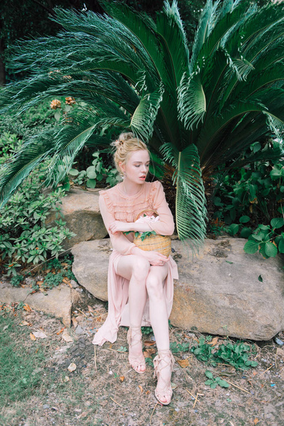 Zmysłowa młoda blondynka piękne słodkie i eleganckie kobieta noszenie różowy siedzi na skale i patrząc w aparat nad tropikalnym zielonym tle palmy dżungli w Chinach. - Zdjęcie, obraz