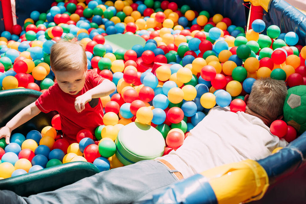 Happy laughing boy 1-2 years old having fun in ball pit on birthday party in kids amusement park and indoor play center. Child playing with colorful balls in playground ball pool. Activity toys for little kid. - Фото, зображення