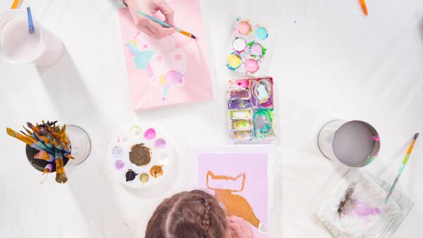 Пласка лежала. Маленька дівчинка малює акриловою фарбою на полотні зі своєю матір'ю для мистецького проекту дистанційного навчання
. - Фото, зображення
