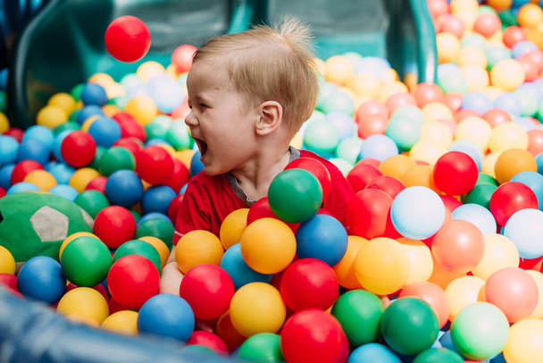 Happy laughing boy 1-2 years old having fun in ball pit on birthday party in kids amusement park and indoor play center. Child playing with colorful balls in playground ball pool. Activity toys for little kid. - Photo, Image