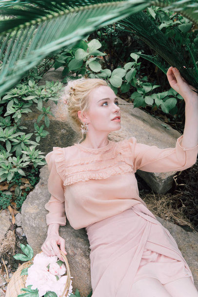 Sensual young blonde beautiful cute and elegant woman wearing pink sitting on the rock and looking into the camera over tropical green jungle palm background in China. - Φωτογραφία, εικόνα
