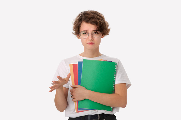 Desapontado jovem estudante em uma camisa branca e óculos segurando pastas coloridas isoladas sobre fundo branco e mostrando gesto so-so. Conceito de educação
 - Foto, Imagem