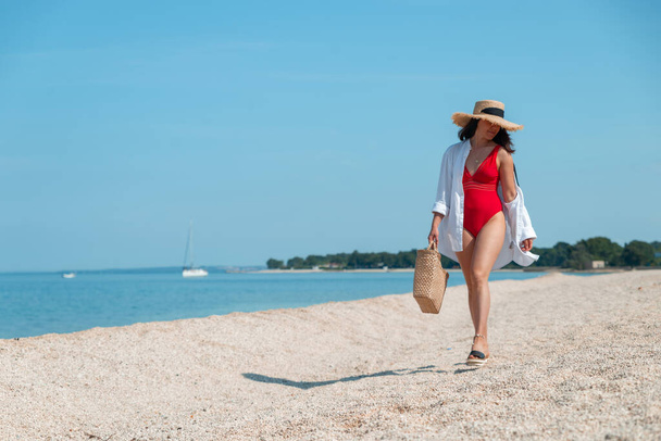 young stunning woman in red swimsuit walking by sea beach. summer outfit - Photo, Image