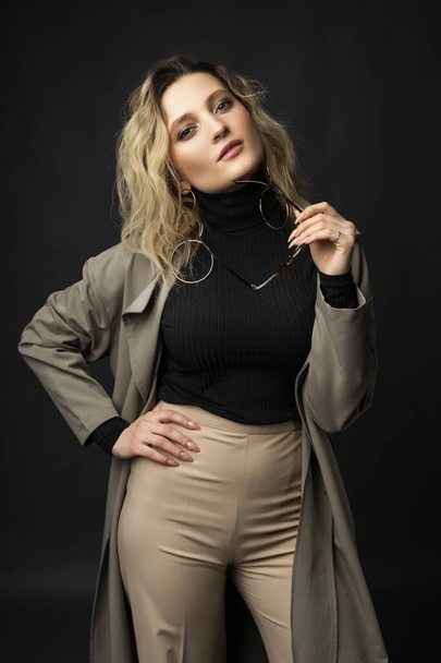 Beautiful girl wearing a casual trendy trousers, turtleneck glasses and raincoat posing on a dark background. Fashionable, advertising, lifestyle and commercial design - Foto, imagen