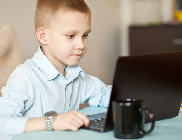 Workspace office desk with laptop and cup of coffee. Little cute business boy sitting at table and looking on a important documents. Kid learning online. Distance learning online education. - 写真・画像
