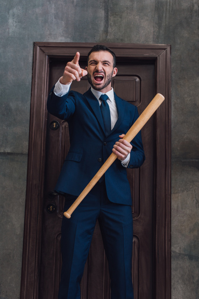 Angry collector with baseball bat pointing with finger and shouting near door in room - Fotoğraf, Görsel