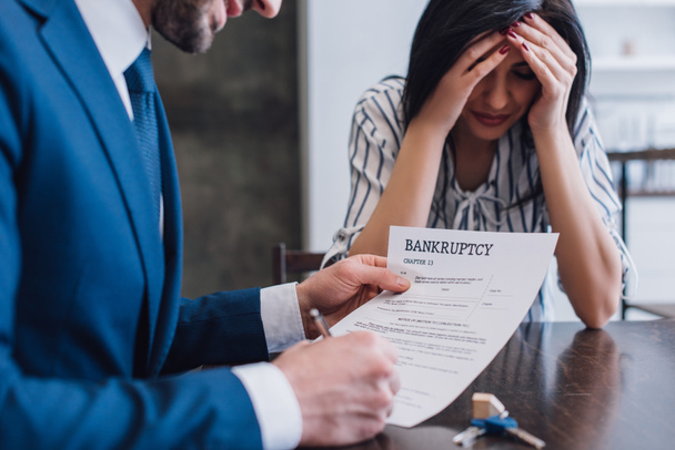 Selective focus of upset woman covering face near collector writing in document with bankruptcy lettering at table - Photo, Image