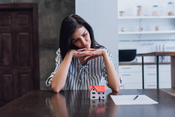 Zdenerwowana i troskliwa kobieta z zaciśniętymi dłońmi przy stole z modelką domu w pobliżu dokumentu w pokoju - Zdjęcie, obraz