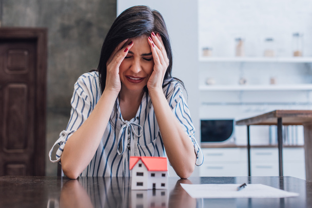 Расстроенная женщина плачет за столом с моделью дома рядом с документом в комнате
 - Фото, изображение