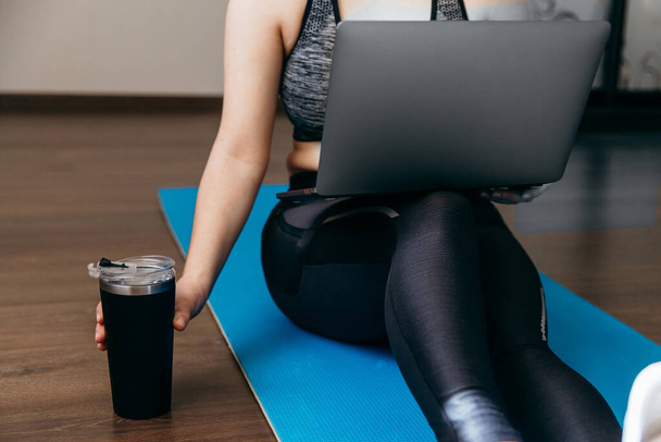 A sporty woman in sportswear is sitting on the floor with dumbbells and a protein shake or a bottle of water and is using a laptop at home in the living room. Sport and recreation concept. - Foto, Imagem