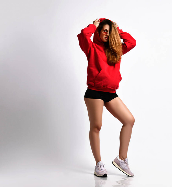 Pregnant lady in black shorts, red hoodie and sunglasses, pink sneakers. She is putting on a hood, isolated on white. Full-length - Valokuva, kuva