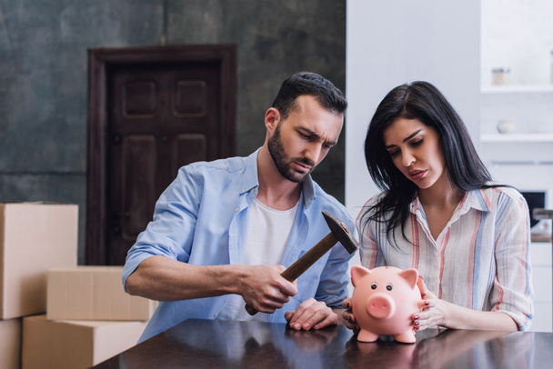 Woman with upset man holding hammer above piggy bank at table in room - Photo, Image