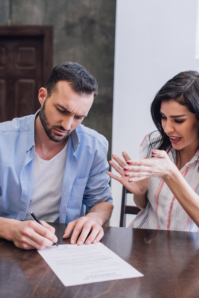 Stressed woman near man writing in document at table in room - Photo, Image