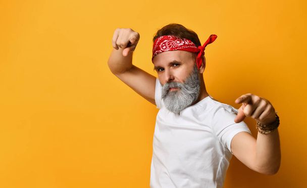 Elderly man in red bandana, white t-shirt and bracelet. He pointing at you by forefingers while posing against orange background - Фото, изображение