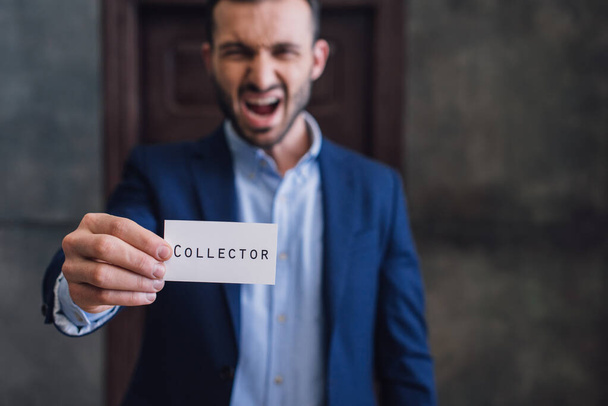 Enfoque selectivo de coleccionista enojado gritando y mostrando tarjeta con letras de coleccionista en la habitación
 - Foto, Imagen