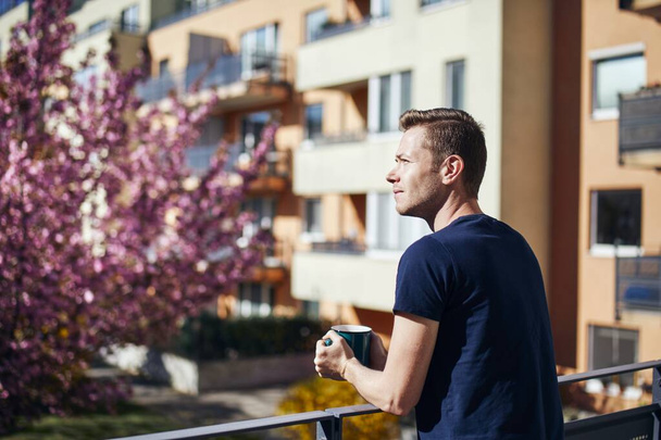 Spring morning at home. Young man with cup tea or coffee on balcony. - Photo, Image