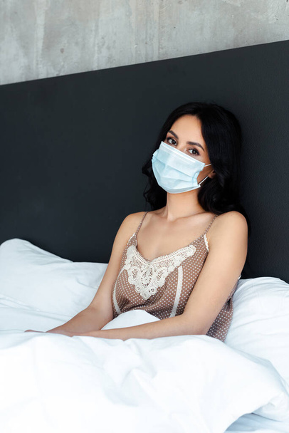 aantrekkelijke vrouw in medisch masker zitten in bed thuis op zelf isolatie - Foto, afbeelding