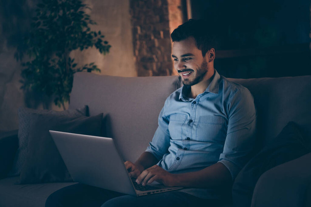 Profile side view portrait of his he nice attractive cheerful cheery brunet guy sitting on divan working remotely typing e-mail letter at modern industrial loft style interior dark living-room office - Zdjęcie, obraz