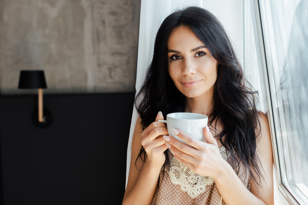 atractiva mujer joven sosteniendo taza de café cerca de la ventana en casa
 - Foto, Imagen