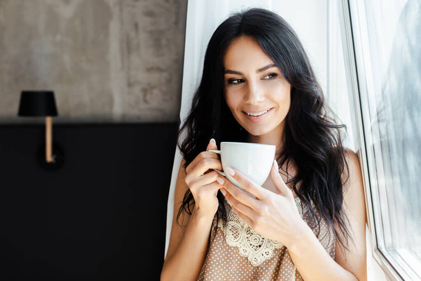 smiling young woman holding cup of coffee and looking through window  - Foto, imagen
