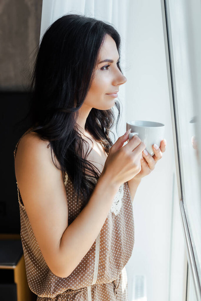 beautiful girl holding cup of coffee and looking through window  - Photo, Image