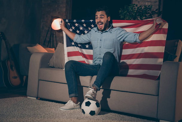 Portrait of his he nice attractive glad crazy overjoyed cheery cheerful brunet guy sitting on divan watching TV sport show holding in hand USA flag at industrial loft style interior house - Fotografie, Obrázek