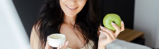 cropped view of smiling woman holding apple and plastic container with cosmetic cream in bedroom, horizontal image - Photo, Image