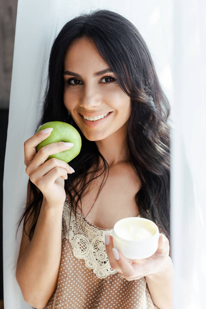 θετική γυναίκα κρατώντας πράσινο μήλο και πλαστικό δοχείο με κρέμα προσώπου  - Φωτογραφία, εικόνα