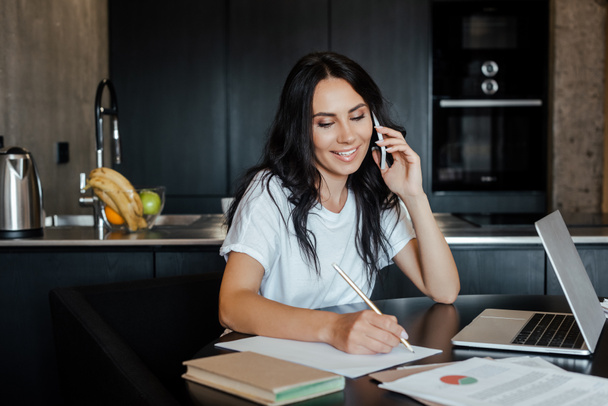 χαμογελαστή γυναίκα που μιλάει στο smartphone και εργάζεται με laptop και επιχειρηματικά έγγραφα στην κουζίνα κατά τη διάρκεια της απομόνωσης - Φωτογραφία, εικόνα