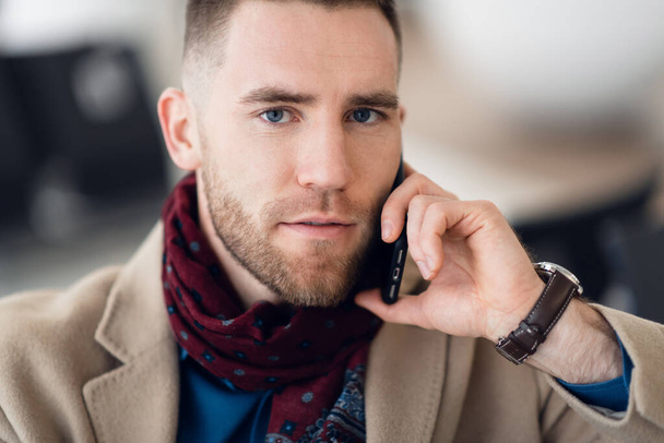 Attractive young adult man in stylish scarf and coat sitting inside airport terminal waiting area. He looking at cellular mobile phone screen and smiling - Foto, Imagem