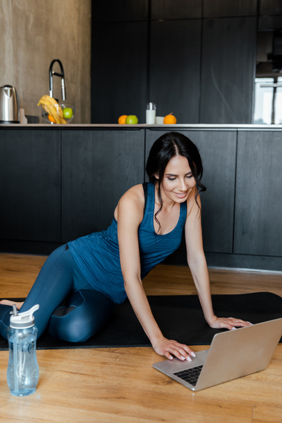 smiling beautiful sportswoman sitting on fitness mat with bottle of water and laptop - Foto, imagen