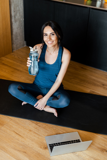 smiling sportive woman holding bottle of water while sitting on fitness mat with laptop - 写真・画像