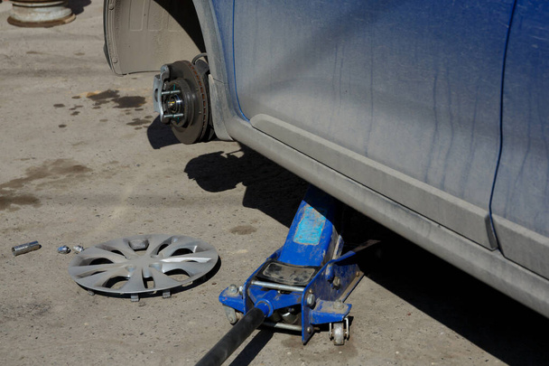 A blue dirty car is standing on a jack without a wheel. Seasonal tire change. Brake disc and pad - Fotografie, Obrázek