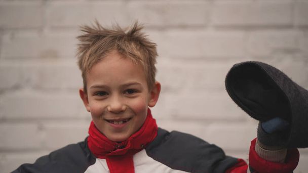 Portrét malého roztomilého chlapce v bundě s červeným límečkem s dětskou čepicí v ruce. Dítě se usmívá a zvednutou rukou mává kloboukem - Fotografie, Obrázek