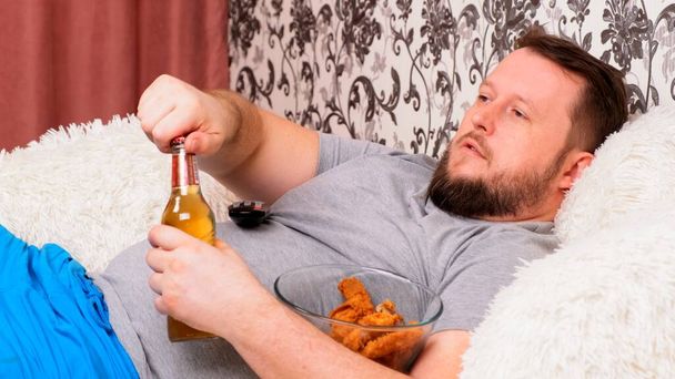 бородатый толстяк с большим животом лежит на диване с фаст-фудом, открывает пиво и скучает по телевизору с пультом дистанционного управления в руке, крупным планом
. - Фото, изображение