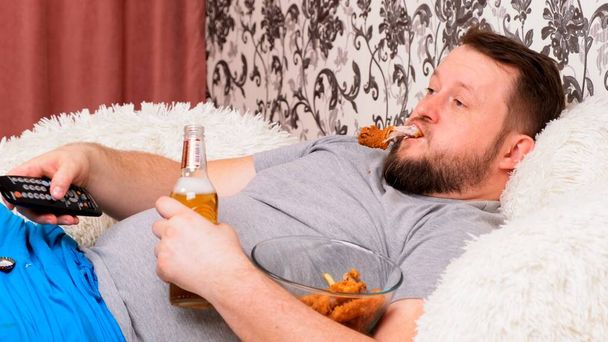大きな腹を持つ髭の太った男は、ファーストフードとソファの上に横たわっています,ビールを飲み、彼の手のリモコンでテレビを欠場,クローズアップ. - 写真・画像