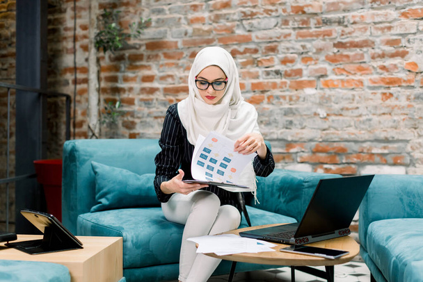Чудова успішна арабська бізнес-жінка сидить на блакитному дивані перед цегляною стіною і працює з інформацією про проекти на папері та ноутбуці. Мусульманка працює в сучасному офісі. - Фото, зображення