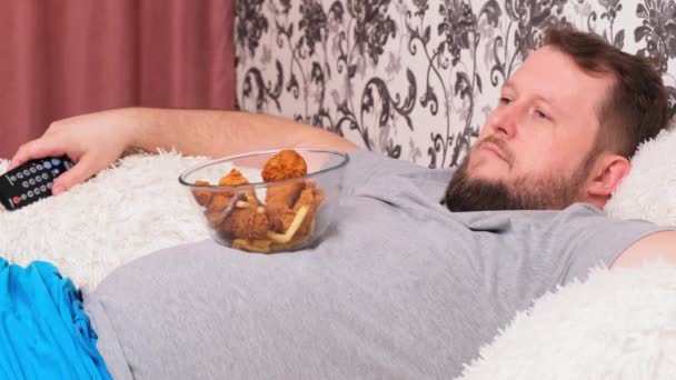 Koca göbekli şişman bir adam elinde fast food ile kanepede yatıyor ve elinde uzaktan kumandayla televizyonun önünde sıkılıyor.. - Video, Çekim