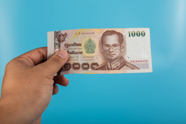 A mans hand is holding one 1000 bank notes on a blue background. Concept Thai baht currency banknote - Photo, Image