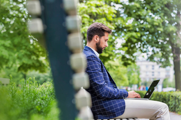 Portrait of young attractive businessman sitting on bench while working on his laptop - 写真・画像