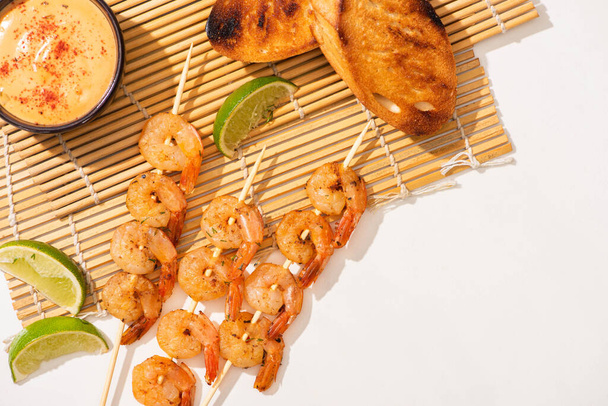 top view of prawns on skewers with toasts, lime and sauce on bamboo mat on white background - Foto, afbeelding
