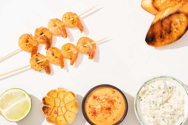 top view of delicious fried prawns on skewers with grilled bread, garlic, lime and sauces on white background - Photo, image