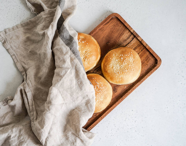 Group crisp fresh homemade buns with sesame on wood tray under a linen towel on a table. Top view - Photo, Image